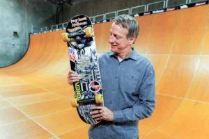 Tony Hawk smiling while holding his Birdhouse Falcon 2 skateboard, the historic deck used to land the first-ever 900 at the 1999 X Games, standing on a vert ramp.