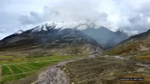 Aerial view of a Himalayan mountain ridge near Shigatse, Tibet, with green fields and rocky terrain in the foreground, snow-capped peaks in the distance