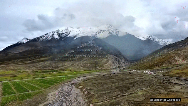 Aerial view of a Himalayan mountain ridge near Shigatse, Tibet, with green fields and rocky terrain in the foreground, snow-capped peaks in the distance