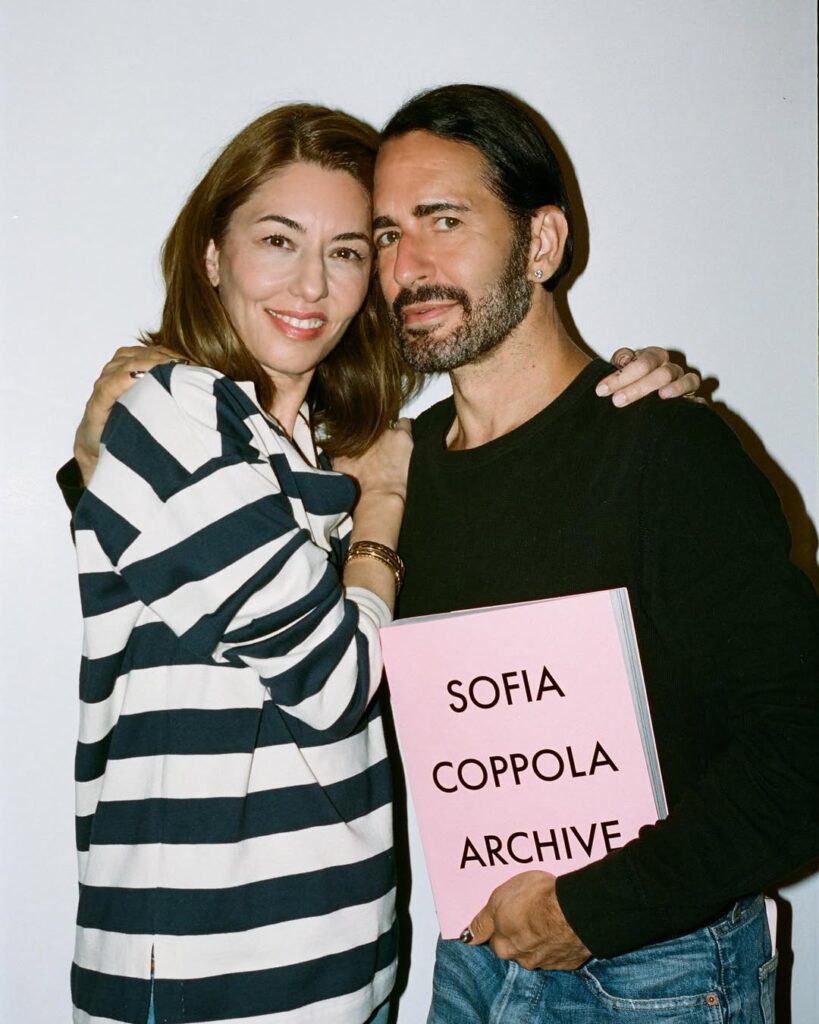 Sofia Coppola and Marc Jacobs posing together, smiling warmly. Coppola wears a striped top while Jacobs holds a pink book titled “Sofia Coppola Archive.”