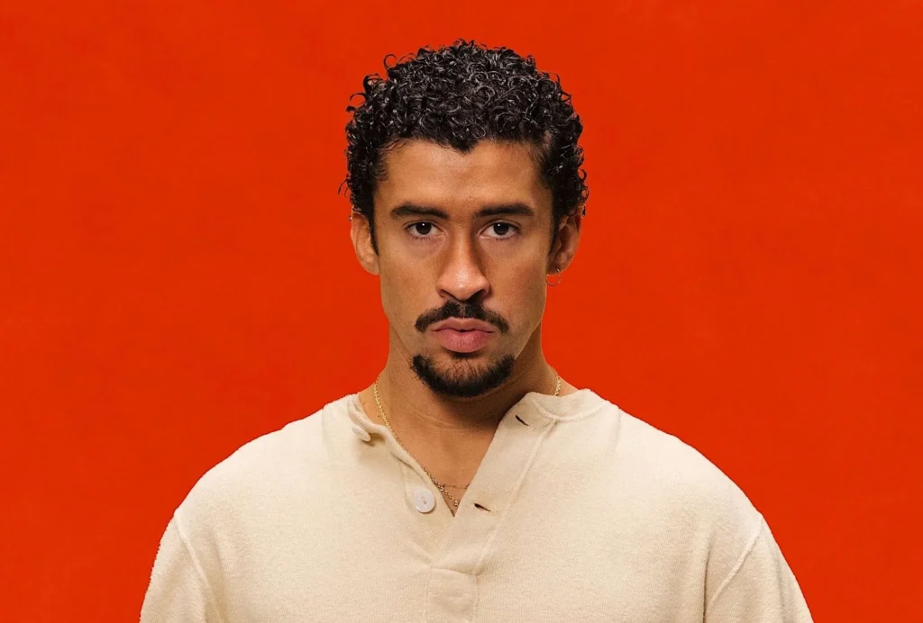Bad Bunny photographed against a solid red background, wearing a cream-colored buttoned shirt, with a serious expression and short curly hair.