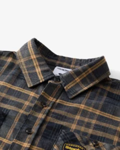 Close-up view of the Staple Pigeon CARTER Flannel shirt in navy and tan plaid, showing button closure, collar detail, and woven “Hardware Staple” patch on the chest pocket.