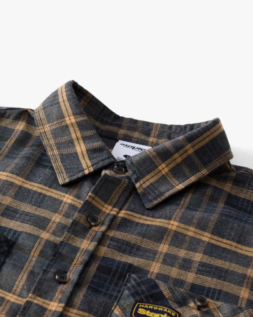 Close-up view of the Staple Pigeon CARTER Flannel shirt in navy and tan plaid, showing button closure, collar detail, and woven “Hardware Staple” patch on the chest pocket.