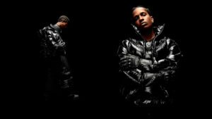 A$AP Rocky wearing a glossy black Moncler puffer jacket from his Moncler Genius collection, photographed against a black background with arms crossed in a bold, high-contrast portrait.