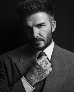 Black and white portrait of David Beckham adjusting his tie while wearing a tailored suit, showcasing mature sophistication and his signature tattoos, part of the Beckham x Boss Fall/Winter 2025 campaign.