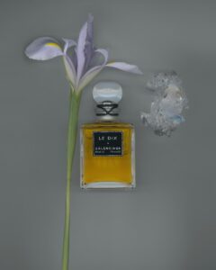 A vintage Balenciaga Le Dix perfume bottle filled with golden liquid, featuring a black label and clear round stopper, styled beside a delicate lavender iris flower and a translucent crystal on a gray background.