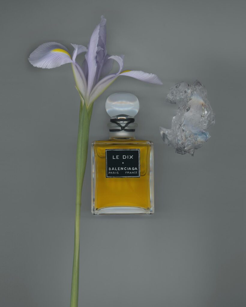 A vintage Balenciaga Le Dix perfume bottle filled with golden liquid, featuring a black label and clear round stopper, styled beside a delicate lavender iris flower and a translucent crystal on a gray background.