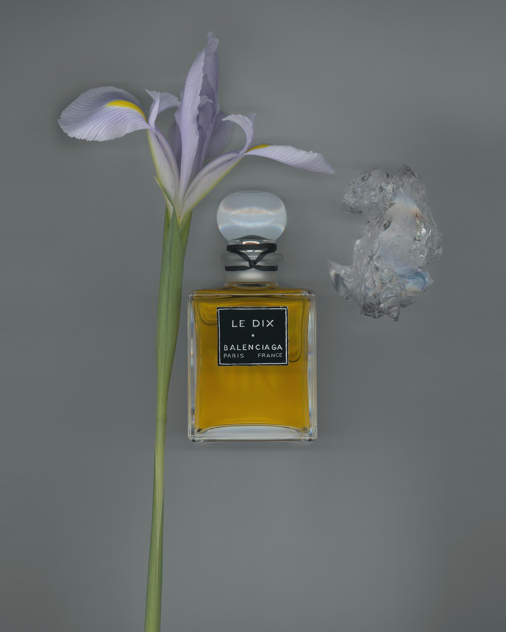 A vintage Balenciaga Le Dix perfume bottle filled with golden liquid, featuring a black label and clear round stopper, styled beside a delicate lavender iris flower and a translucent crystal on a gray background.