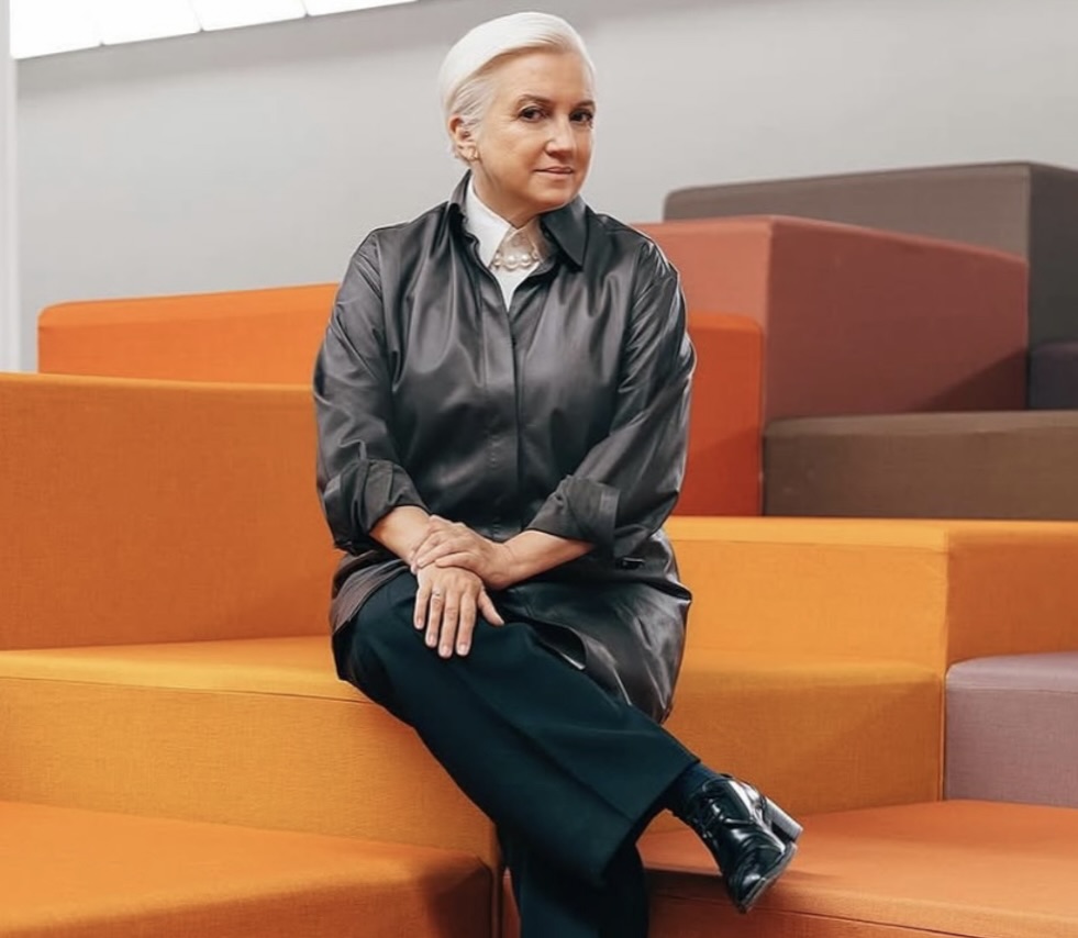 Silvia Venturini Fendi seated on bright orange and brown tiered steps, wearing a dark satin shirt with pearl necklace, black trousers, and polished shoes, as she prepares for her new role as honorary president of Fendi.