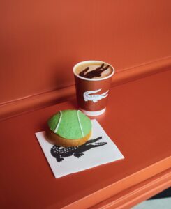 A Lacoste-branded coffee cup topped with a cocoa crocodile motif, paired with a green tennis ball-shaped brioche pastry on a Lacoste napkin, set against a clay-court-inspired red background.