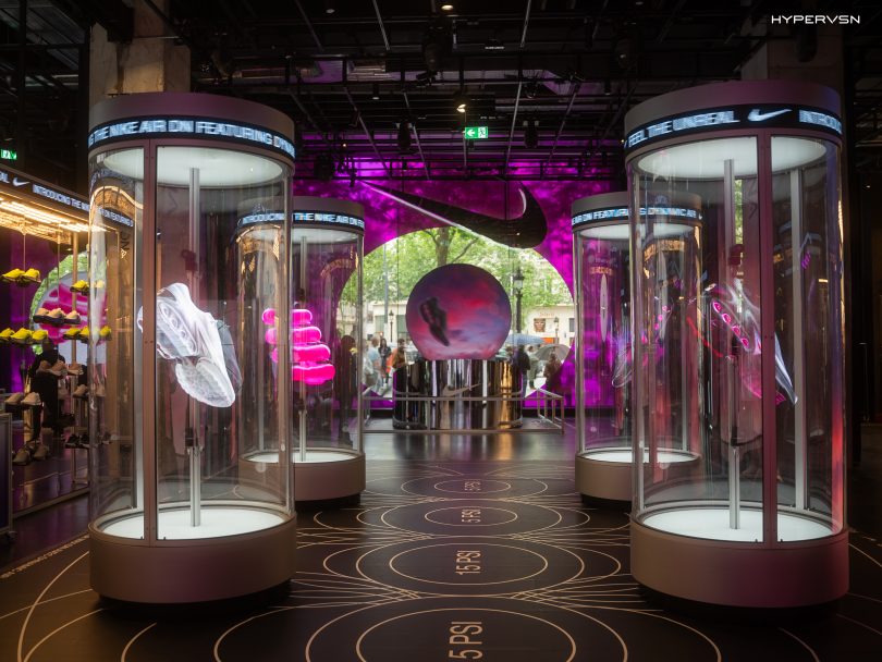 Holographic sneaker displays inside Nike’s House of Innovation in Paris, featuring cylindrical glass pods projecting floating 3D Air Max Dn visuals with vibrant purple lighting and a large Nike logo backdrop.