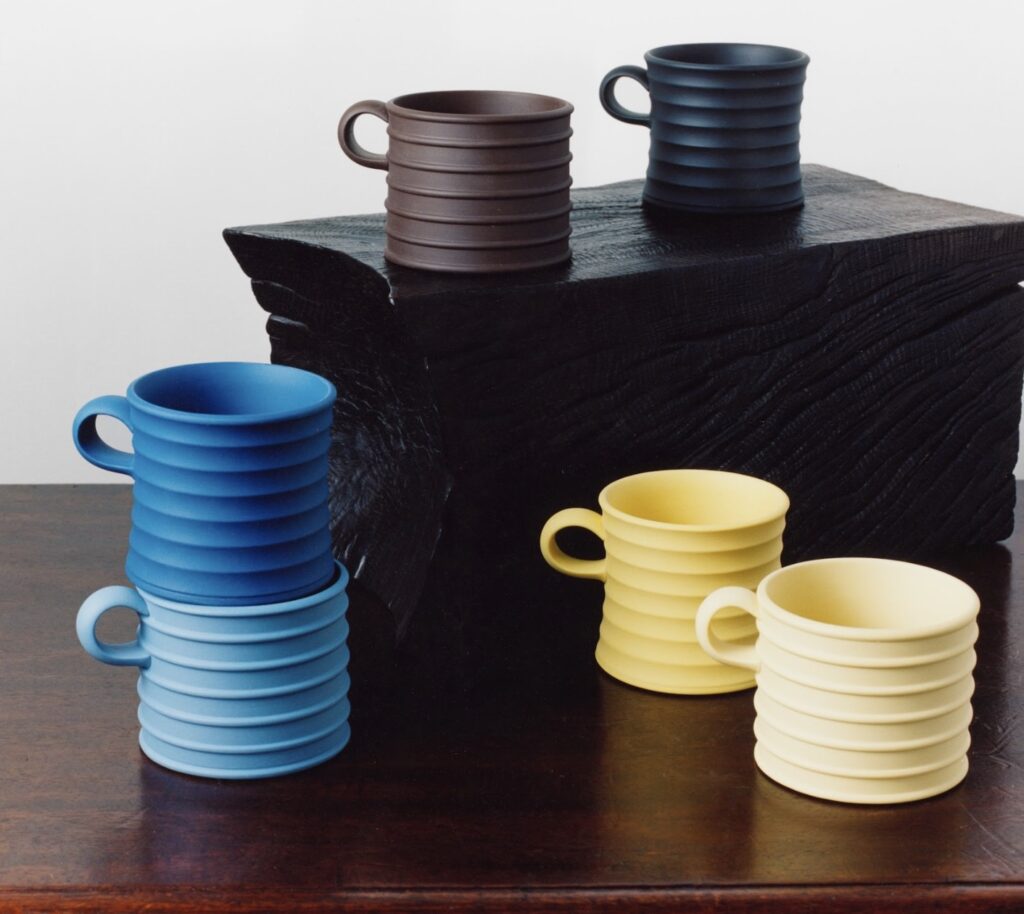 A set of ribbed ceramic mugs from the JW Anderson x Wedgwood collaboration, shown in bold matte colors including chocolate brown, black, bright blue, light blue, and pale yellow, arranged on a dark wooden table and a black textured block.