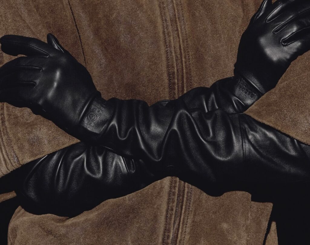 Close-up of crossed arms wearing sleek black leather gloves from the MM6 Maison Margiela x Agnelle collection, styled over a brown shearling coat.
