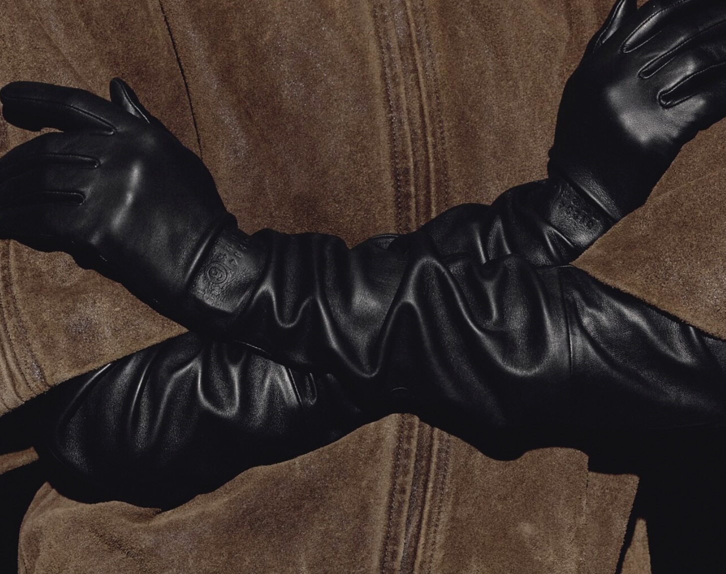 Close-up of crossed arms wearing sleek black leather gloves from the MM6 Maison Margiela x Agnelle collection, styled over a brown shearling coat.
