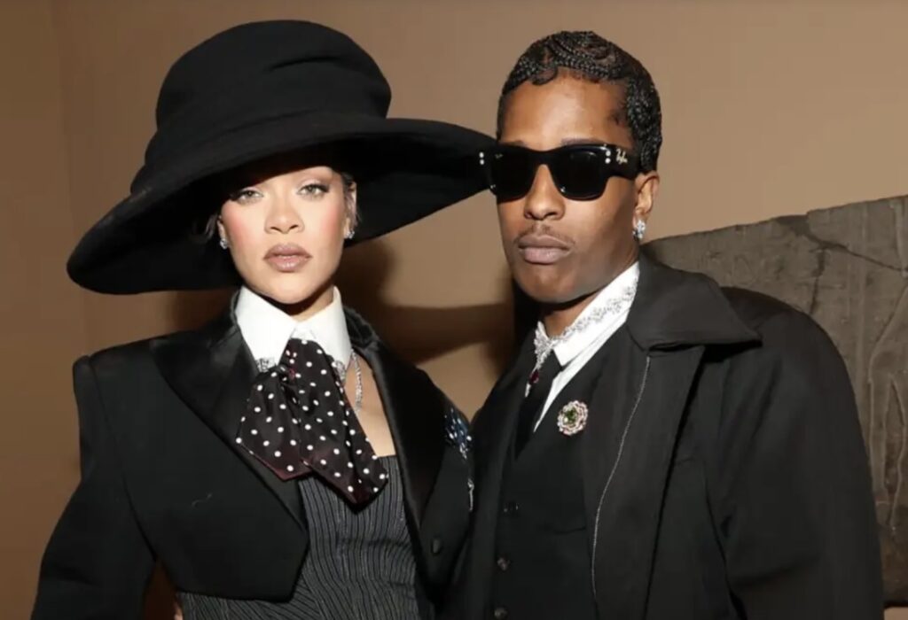 Rihanna and A$AP Rocky pose together in coordinated black outfits. Rihanna wears a dramatic wide-brimmed black hat, a tailored blazer with structured shoulders, a pinstriped vest, and a polka-dot necktie. A$AP Rocky wears dark sunglasses, a black overcoat layered over a suit and white shirt, accessorized with jeweled brooches and earrings.