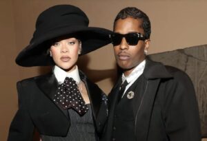 Rihanna and A$AP Rocky pose together in coordinated black outfits. Rihanna wears a dramatic wide-brimmed black hat, a tailored blazer with structured shoulders, a pinstriped vest, and a polka-dot necktie. A$AP Rocky wears dark sunglasses, a black overcoat layered over a suit and white shirt, accessorized with jeweled brooches and earrings.