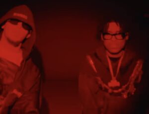 Yan Block and Panda Black in a dimly lit red-tinted scene, wearing hoodies and glasses, captured in a moody, atmospheric moment from their collaboration Talento.