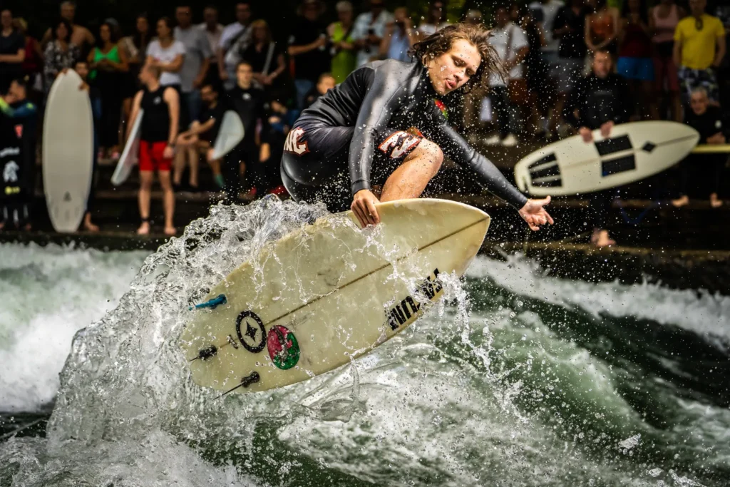Jakob Emilio performing an aerial maneuver on Munich’s Eisbach river wave, with water splashing around and spectators watching from the riverbank.