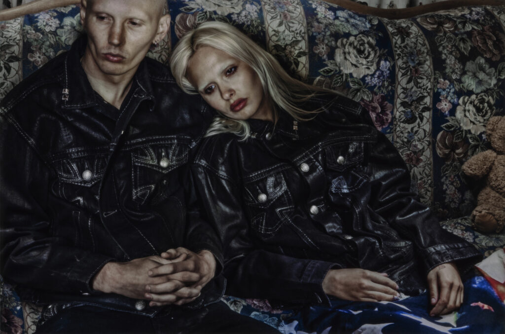 Two models sit closely together on a floral-patterned couch, wearing matching oversized black denim jackets from the Ksubi by Alice Hollywood capsule collection, evoking early-2000s indie sleaze and punk-inspired aesthetics.