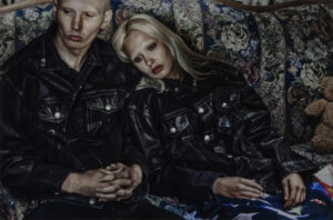 Two models sit closely together on a floral-patterned couch, wearing matching oversized black denim jackets from the Ksubi by Alice Hollywood capsule collection, evoking early-2000s indie sleaze and punk-inspired aesthetics.