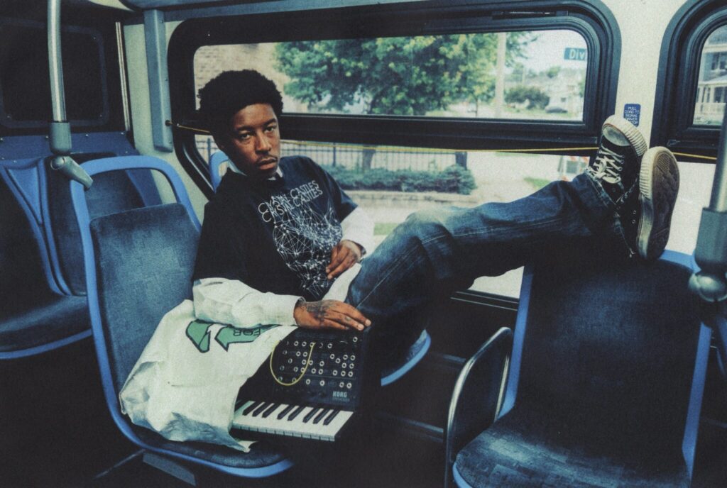 KennyHoopla sitting on a bus with a relaxed pose, wearing a black graphic T-shirt and jeans, resting his feet on the seat in front while holding a small Korg synthesizer keyboard.
