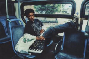 KennyHoopla sitting on a bus with a relaxed pose, wearing a black graphic T-shirt and jeans, resting his feet on the seat in front while holding a small Korg synthesizer keyboard.
