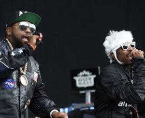 OutKast duo Big Boi and André 3000 performing together on stage; Big Boi in a NASA patch jacket and cap with gold chains, André 3000 in a furry white hat, sunglasses, and dark jacket, both holding microphones mid-performance.