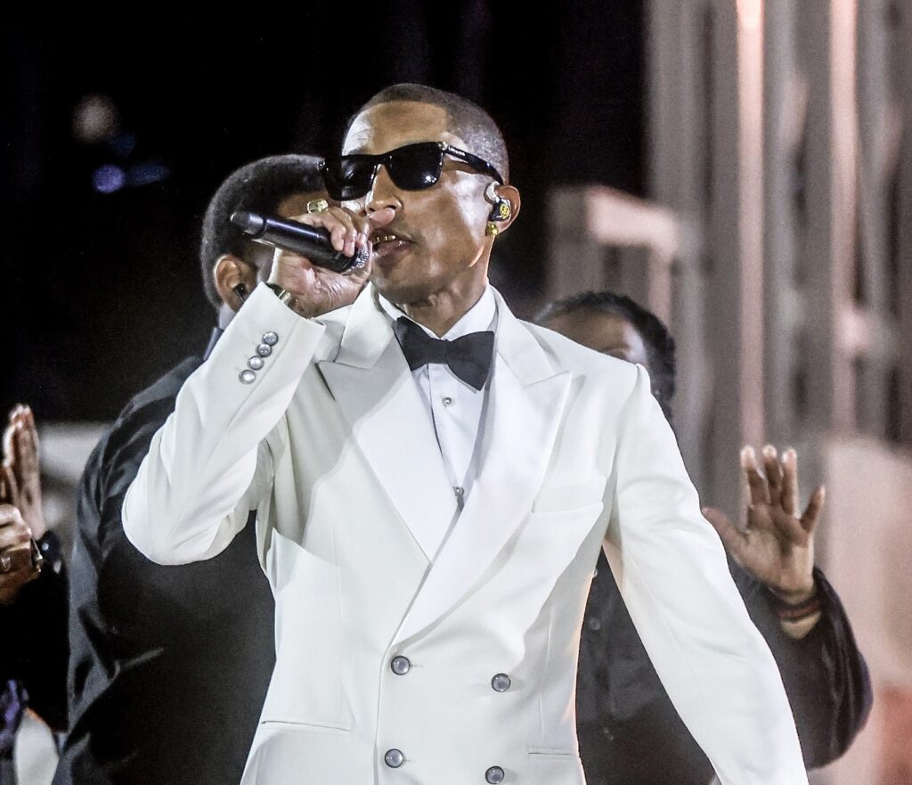 Pharrell Williams performing at the ‘Grace for the World’ concert in Vatican City, wearing a white double-breasted tuxedo jacket with black bow tie and sunglasses, holding a microphone on stage.