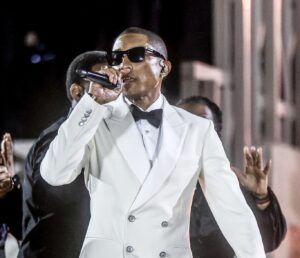 Pharrell Williams performing at the ‘Grace for the World’ concert in Vatican City, wearing a white double-breasted tuxedo jacket with black bow tie and sunglasses, holding a microphone on stage.