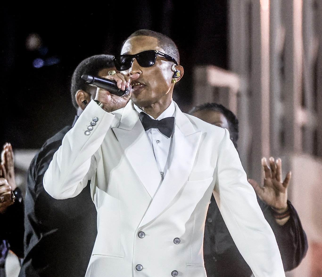 Pharrell Williams performing at the ‘Grace for the World’ concert in Vatican City, wearing a white double-breasted tuxedo jacket with black bow tie and sunglasses, holding a microphone on stage.