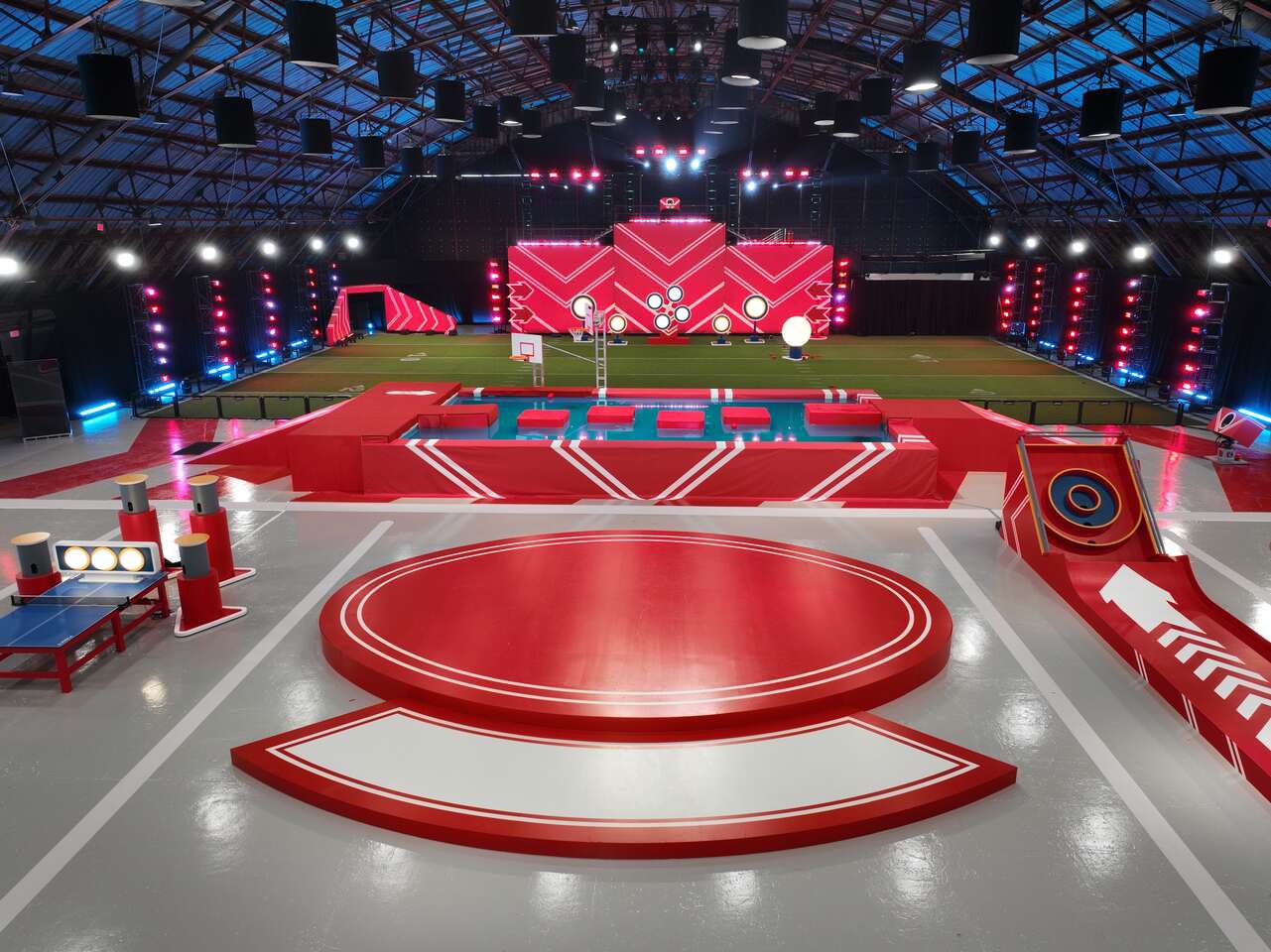 Play it Forward game show set featuring bright red obstacle courses, splash challenges, and stage lighting inside a large arena.