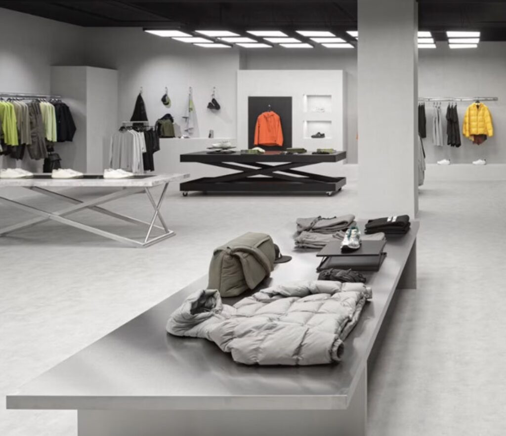 Interior view of Zara’s Rotterdam flagship designed by Random Studio, featuring metallic display tables, geometric blocks, and muted natural tones with overhead light panels illuminating jackets, shirts, and accessories.