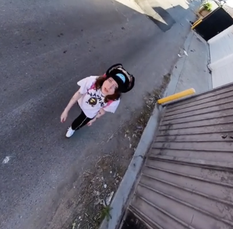 Cr1tter wearing a pink graphic t-shirt, dark pants, and a furry hat looks up toward the camera while standing on an empty street beside a corrugated metal wall.