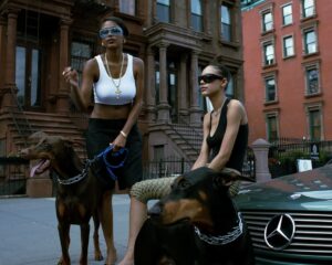 Two women in stylish streetwear pose with Doberman dogs in front of brownstone buildings; one wears a white crop top and gold chain with blue sunglasses, the other sits on a Mercedes car hood in a black tank top and dark sunglasses.