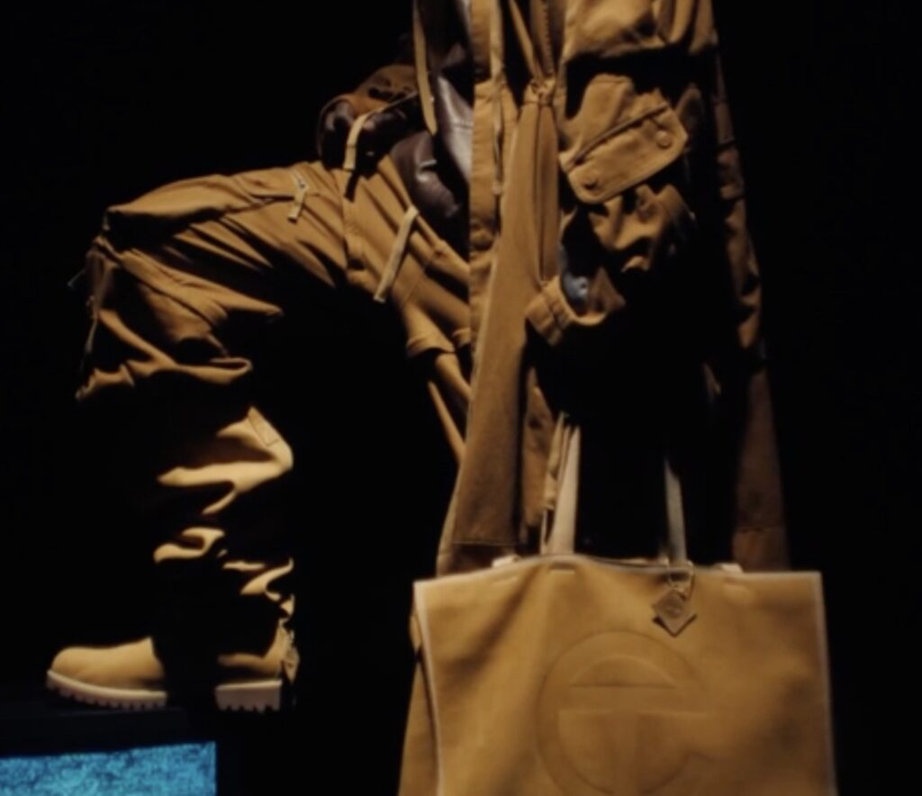 Close-up of Timberland × TELFAR collaboration featuring wheat-colored Timberland boots, utility-style cargo pants, and a large TELFAR Shopping Bag in matching nubuck leather, photographed in dramatic low light.