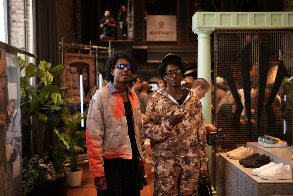 Two men wearing HÉLÀ Code outfits at the NYFW showcase, one in a grey-red jacket with sunglasses and the other in a camouflage tracksuit, standing in front of sneaker displays and brand signage.