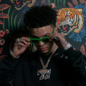 Lil Mosey wearing dark streetwear with layered diamond chains and rings, adjusting bright green sunglasses against a patterned backdrop featuring tigers and floral designs.