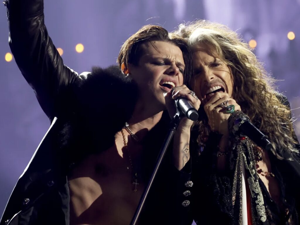 Yungblud and Steven Tyler singing passionately together on stage, sharing a microphone under dramatic concert lighting during their performance of “My Only Angel.