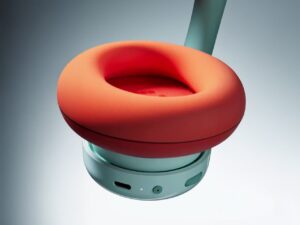 Close-up of the CMF Headphone Pro ear cup, showing a bold orange cushion with soft fabric texture, attached to a mint green frame featuring a USB-C charging port, status light, and control button.