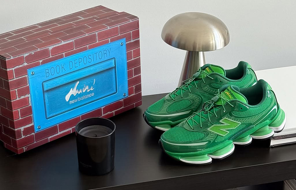 Pair of green Aminé x New Balance ABZORB 2000 sneakers displayed on a black table beside a custom brick-patterned shoebox labeled 'Book Depository,' with a candle and metallic lamp in the background.