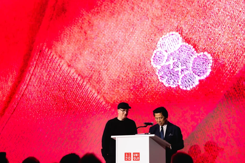 KAWS and UNIQLO executives stand at a podium during the Artist in Residence announcement at MoMA, with a large red backdrop featuring a KAWS Companion motif.
