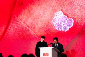 KAWS and UNIQLO executives stand at a podium during the Artist in Residence announcement at MoMA, with a large red backdrop featuring a KAWS Companion motif.