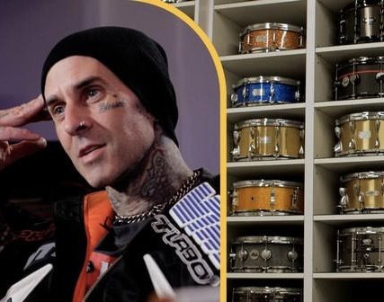 Travis Barker wearing a black beanie and jacket, seated in his studio next to a large shelving unit filled with a colorful collection of snare drums.