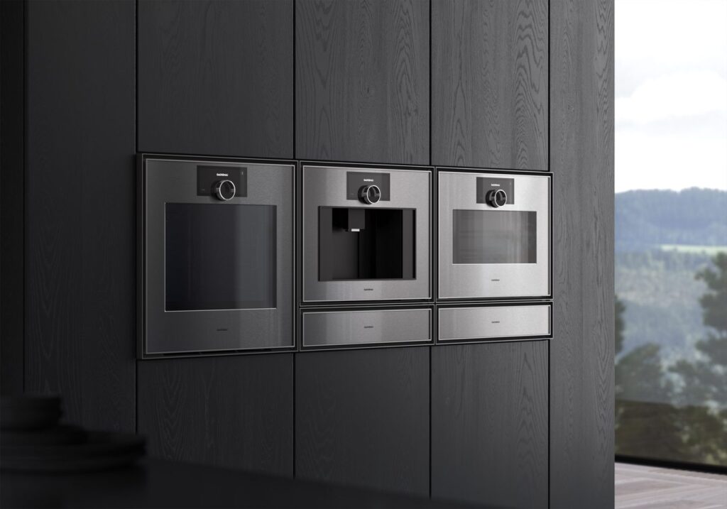 Three Gaggenau Expressive Series built-in appliances—two ovens and a coffee machine—in brushed stainless steel with chamfered edges, seamlessly integrated into a dark wood modern kitchen interior with a view of nature outside.
