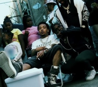 G Herbo sitting casually in a street setting with friends and children around him, wearing a white Satoshi T-shirt and sneakers, embodying a relaxed yet gritty urban atmosphere.