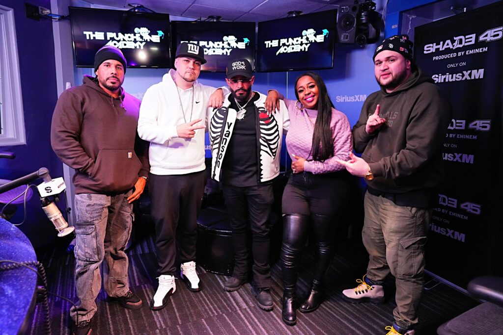 The Punchline Academy show. They stand together in front of screens displaying the Punchline Academy logo, with microphones and studio equipment visible in the room.