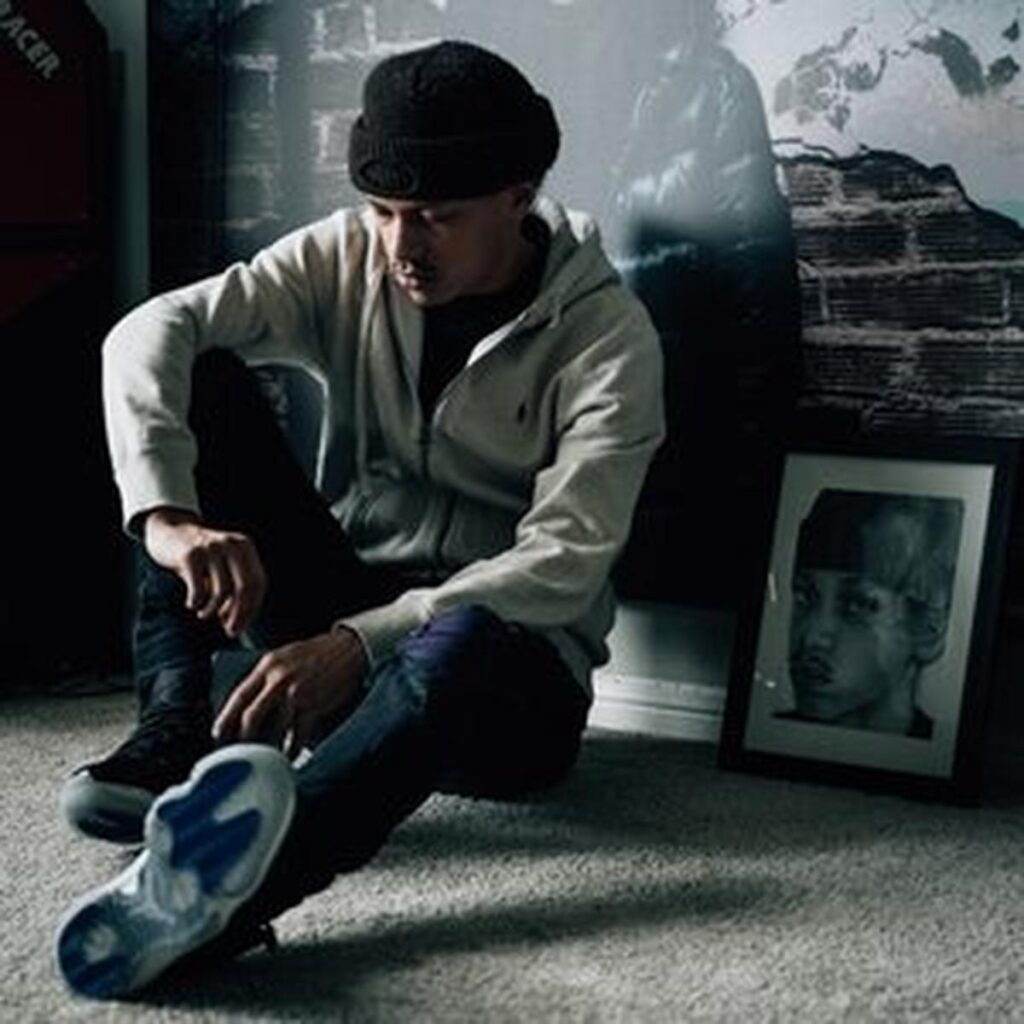Jace, wearing a black beanie, light zip-up hoodie, and dark jeans sits on the floor in a dimly lit room with one knee raised, beside a framed black-and-white portrait, creating a moody, introspective scene.
