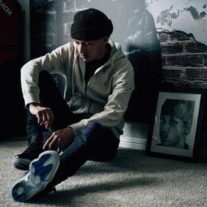 Jace, wearing a black beanie, light zip-up hoodie, and dark jeans sits on the floor in a dimly lit room with one knee raised, beside a framed black-and-white portrait, creating a moody, introspective scene.