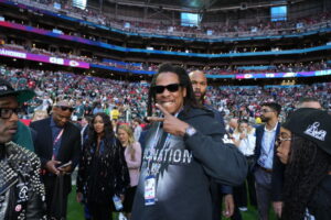 Jay-Z smiling and flashing a peace sign on the field before a major football game, surrounded by Roc Nation affiliates and fans in the stadium background.