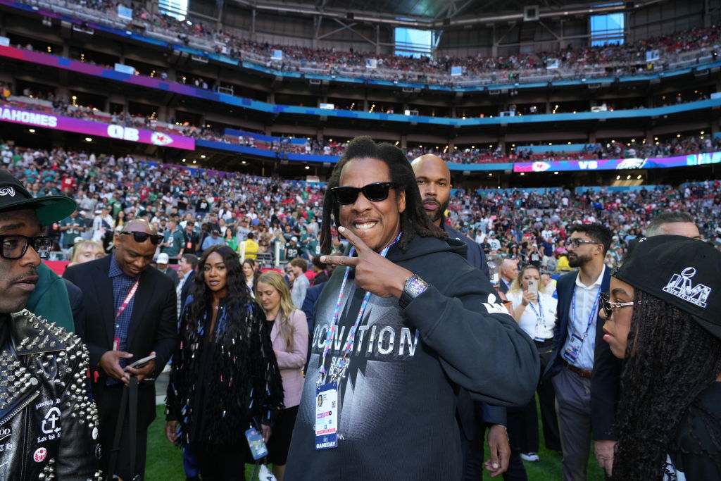 Jay-Z smiling and flashing a peace sign on the field before a major football game, surrounded by Roc Nation affiliates and fans in the stadium background.